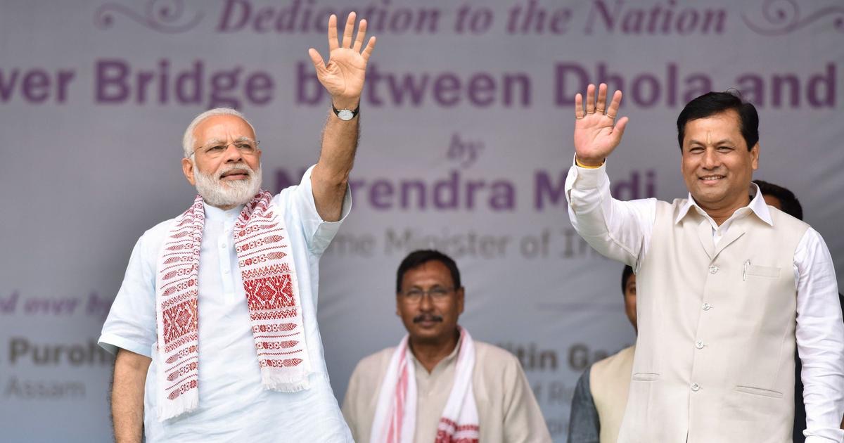 Prime Minister Narendra Modi and Sarbananda Sonowal wave from a stage whose backdrop reads 'Dedication to the Nation — River Bridge between Dhola and Sadiya'
