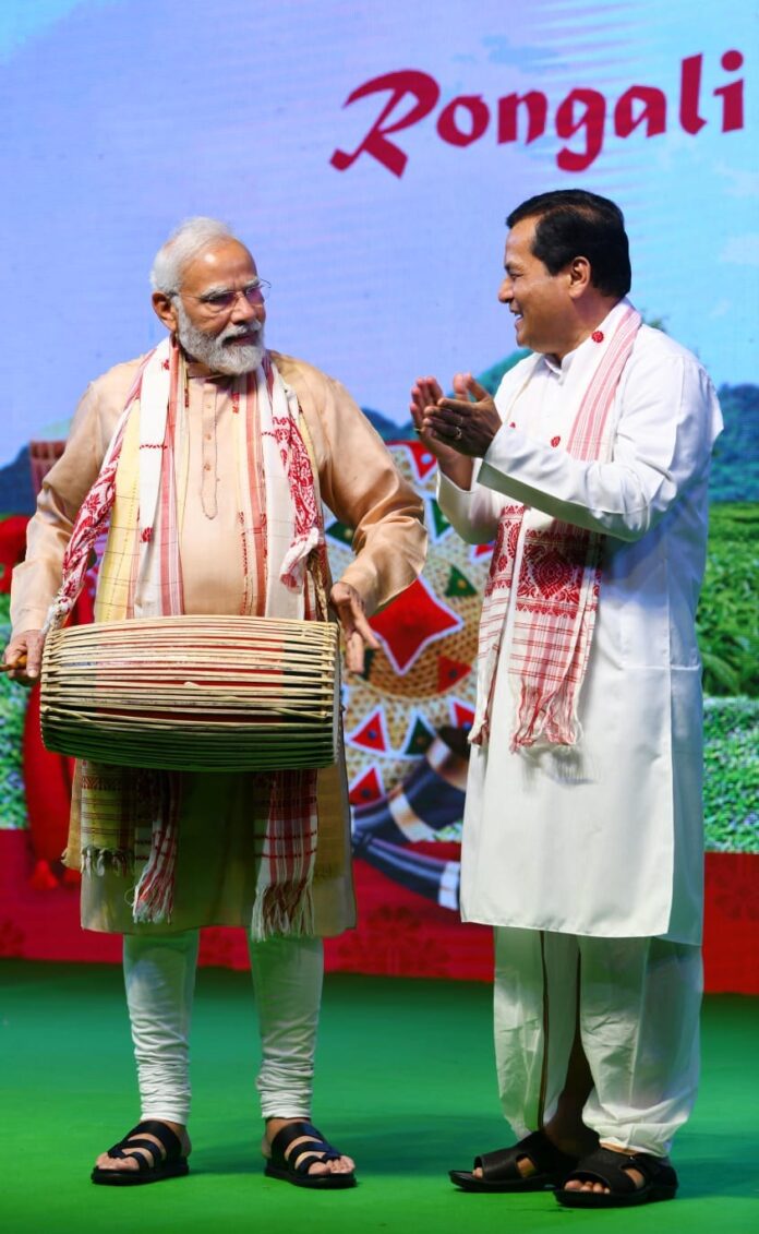 Sarbananda Sonowal, in a white dhoti-kurta and gamosa, applauds as Prime Minister Narendra Modi plays a dhol on a stage decorated for Rongali Bihu