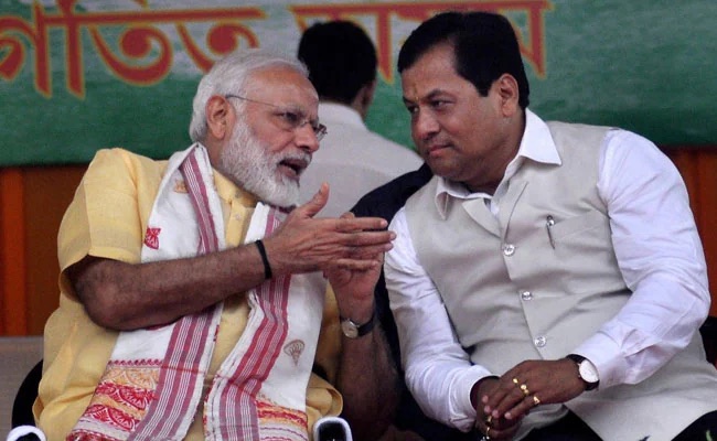 Prime Minister Narendra Modi speaks to Sarbananda Sonowal, seated beside him on a stage in Assam
