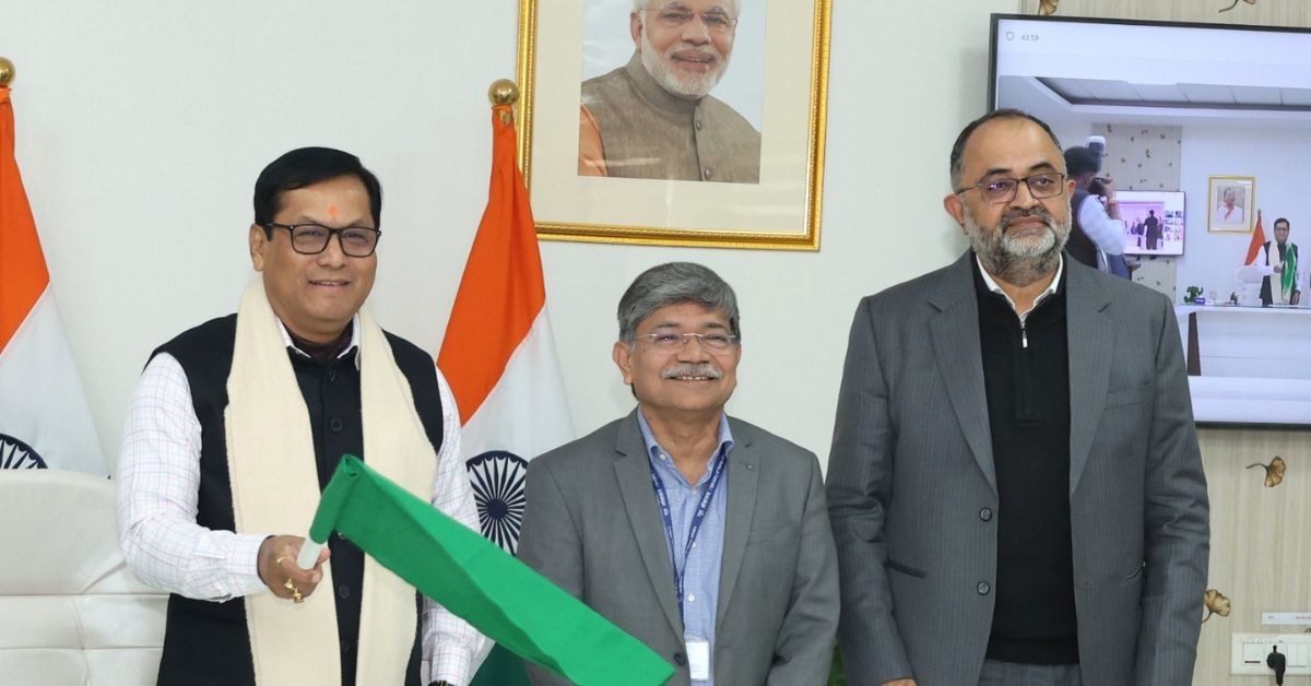 Sarbananda Sonowal waves a green flag at a flag-off ceremony, standing with two officials beside Indian flags