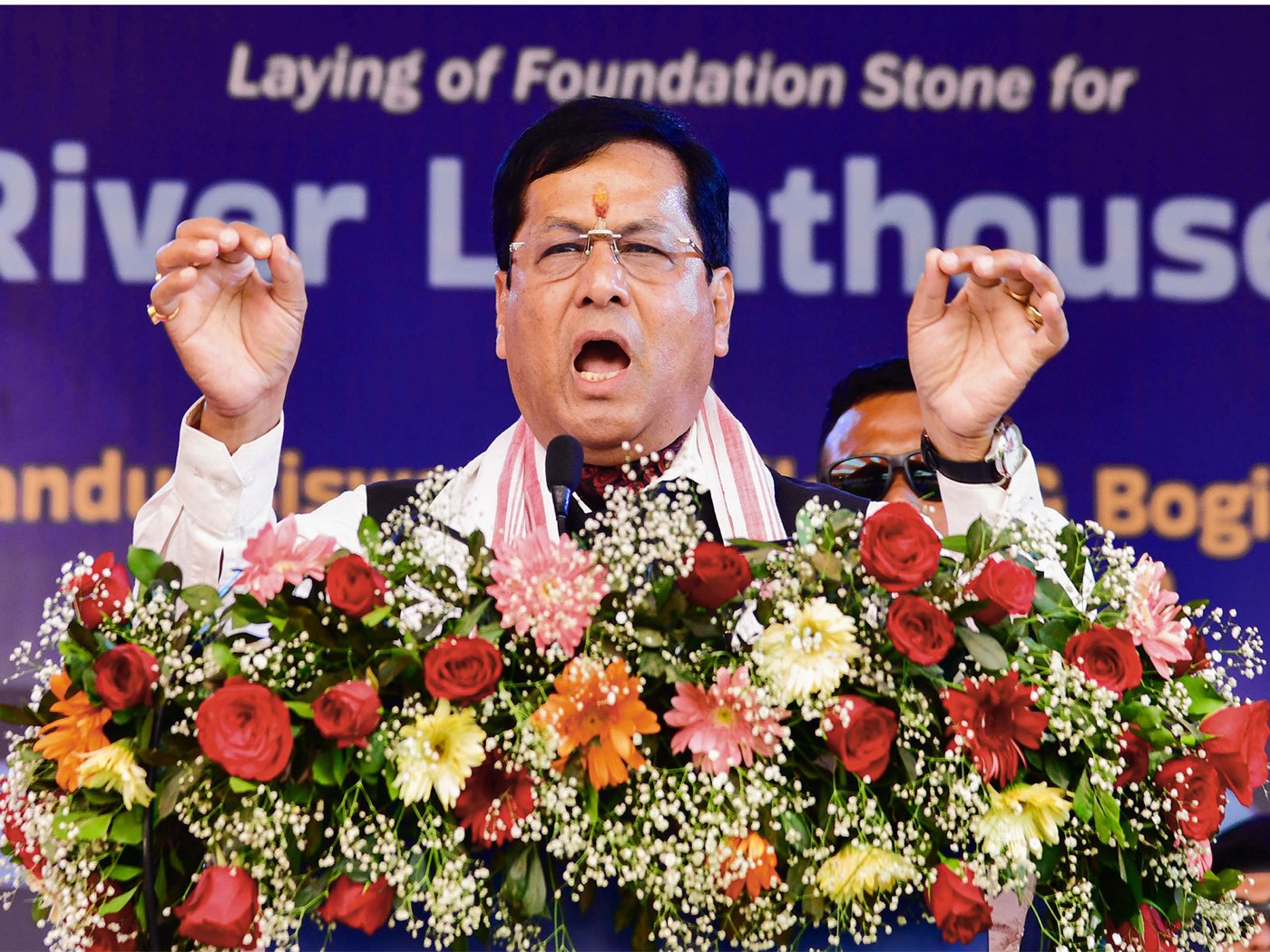 Sarbananda Sonowal, wearing glasses and a gamosa, speaks at a podium covered in flowers, in front of a banner reading 'Laying of Foundation Stone for River Lighthouse'