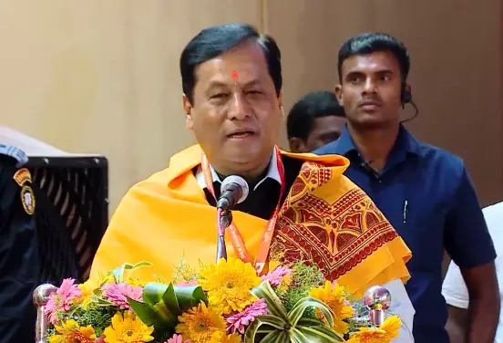 Sarbananda Sonowal, wearing a saffron shawl with a red Assamese motif, speaks into a microphone behind a floral arrangement