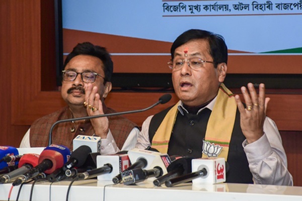 Sarbananda Sonowal, in a black waistcoat and gamosa, speaks at a press conference behind a row of broadcasters' microphones