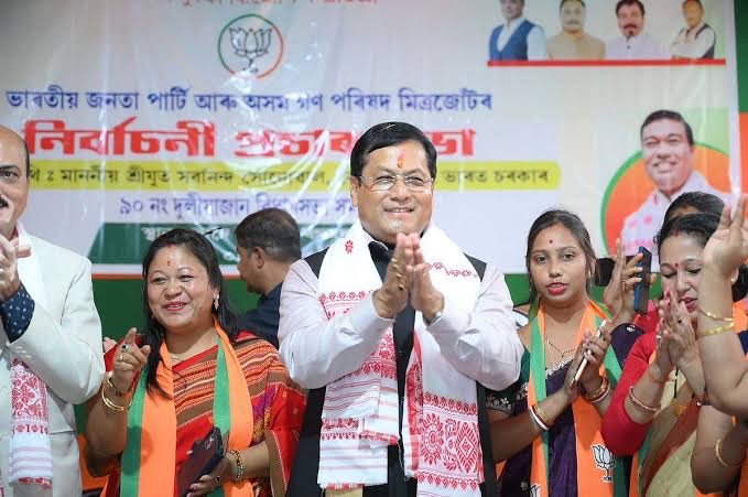 Sarbananda Sonowal greets an audience with folded hands at a meeting, surrounded by party workers, under an Assamese-language banner