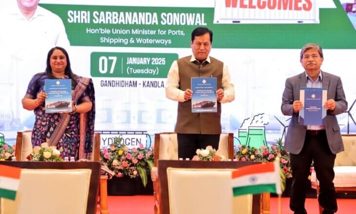 Sarbananda Sonowal and two officials hold up blue publications on a stage whose banner reads 07 January 2025, Gandhidham–Kandla