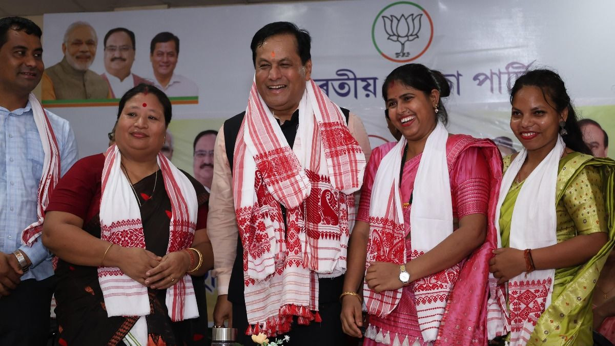 Sarbananda Sonowal, draped in a red-and-white gamosa, laughs with women party workers in front of a party banner in Assamese