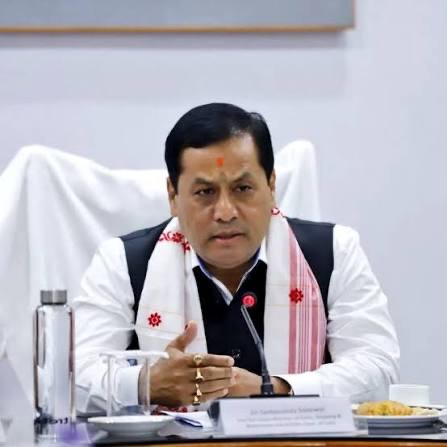 Sarbananda Sonowal, wearing a gamosa, speaks at a meeting table with a nameplate and microphone in front of him