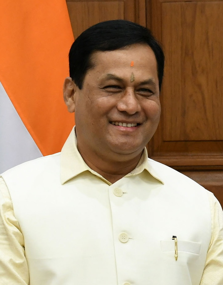 Sarbananda Sonowal, smiling, in a cream Nehru jacket, with the national flag and wood panelling behind him